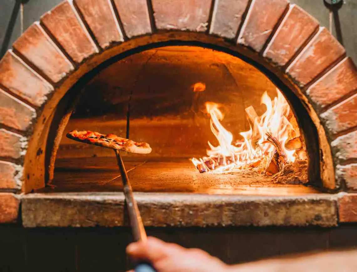 Pizza chef cooking pizza in woodfire oven - 799x610